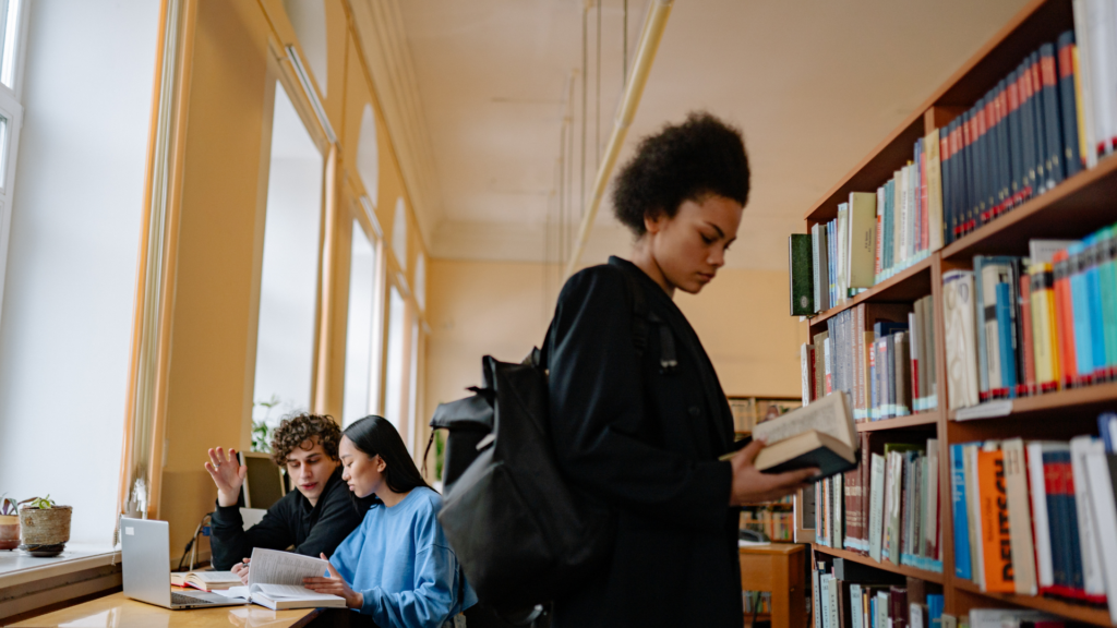 A girl holding a book and reading, with two people chatting in the background inside a library.