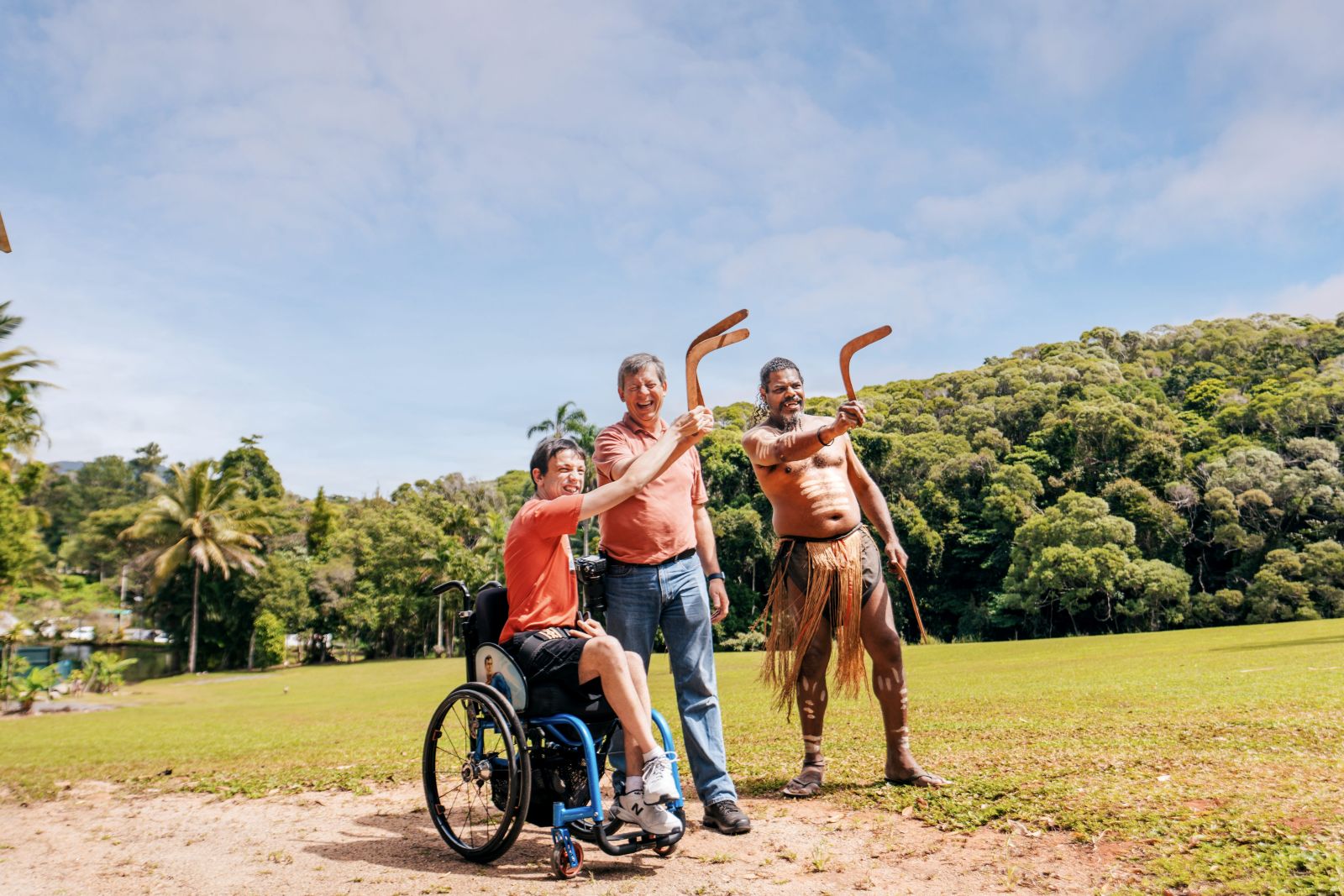 A wheelchair user and his travel companion laugh as they learn boomerang throwing with a Pamagirri guide at Rainforestation Nature Park, rainforest hills behind them.