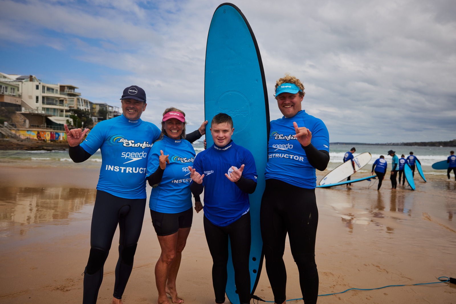 A young surfer with Down syndrome stands with three instructors in front of a surfboard at Bondi Beach, all smiling and giving shaka signs after an adaptive surf lesson.