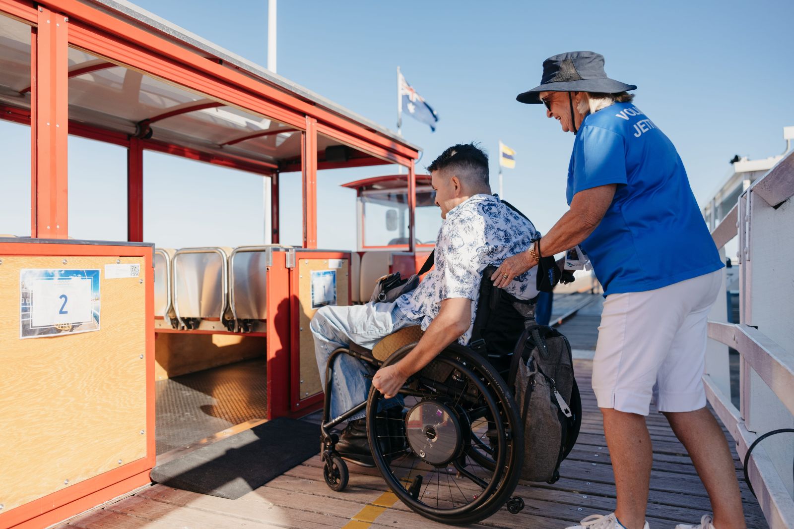 A jetty volunteer assists a wheelchair user boarding the Busselton Jetty train via a level ramp on a sunny day, Australian flag flying above.