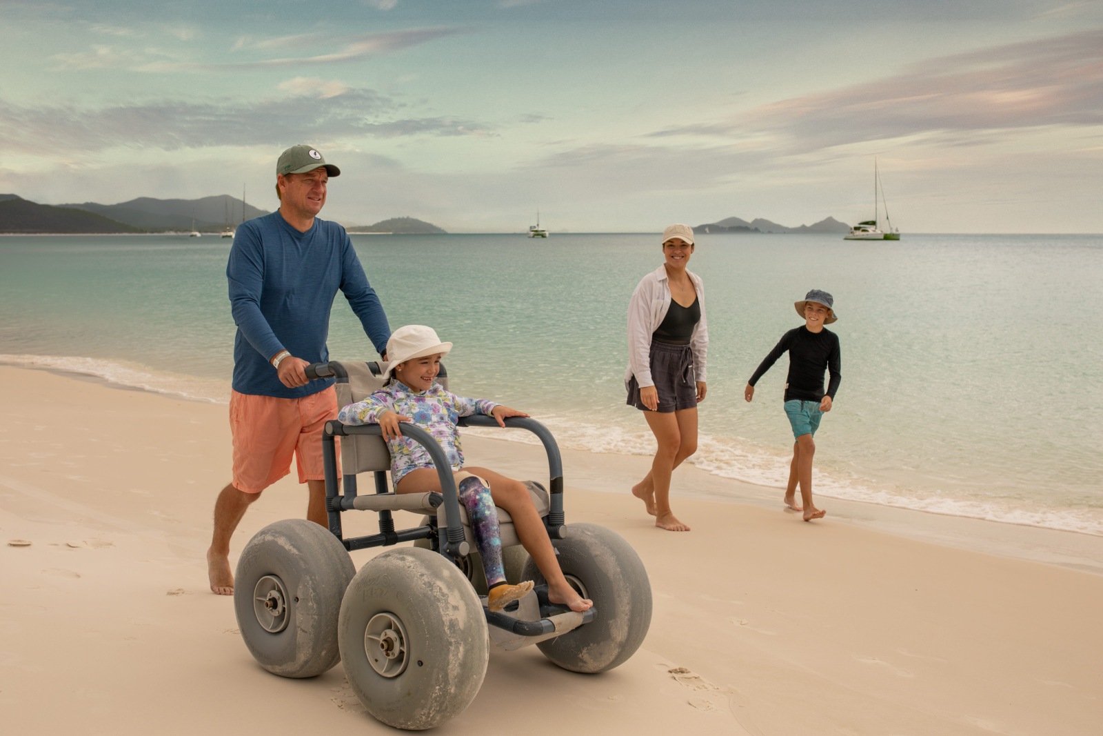 A father pushes his daughter along the shoreline of Whitehaven Beach in a beach wheelchair with balloon tyres, while her mother and brother walk beside them at the water's edge.
