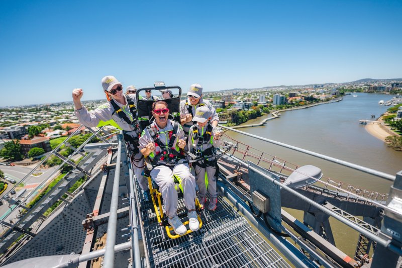 A climber in a motorised stair-climbing wheelchair celebrates with three guides at the summit of Brisbane's Story Bridge, the river and city skyline spread out below.