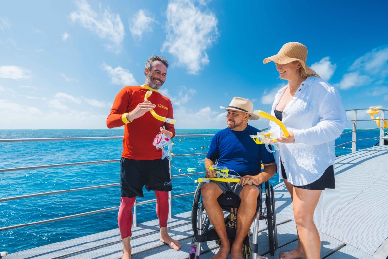 On a reef pontoon deck, a Quicksilver crew member shows snorkelling gear to a wheelchair user and his companion, turquoise Coral Sea water behind them.