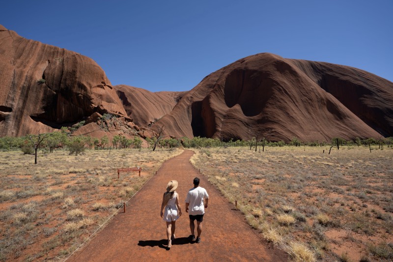 Two travellers walk side by side along the flat red-earth base walk path at Uluru, the rock's curved walls rising against a deep blue sky.