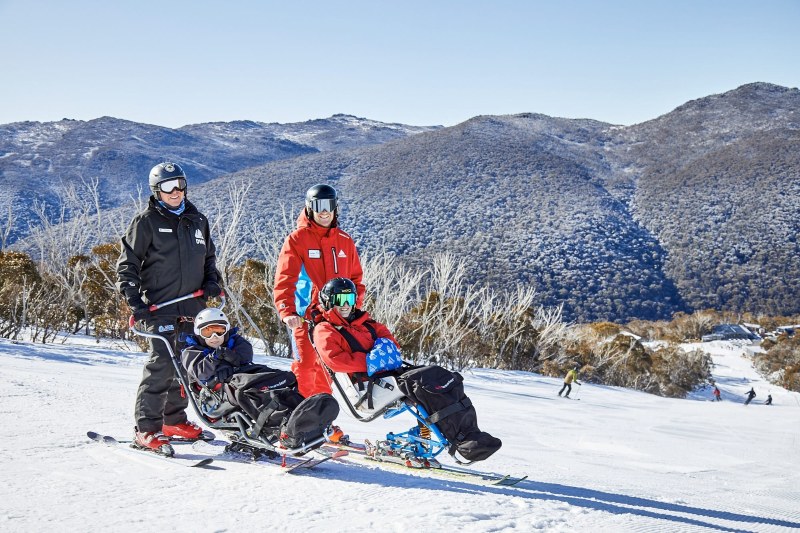 Two sit-skiers and their instructors pause on a groomed run at Thredbo, snow-dusted gums and the Kosciuszko ranges behind them.