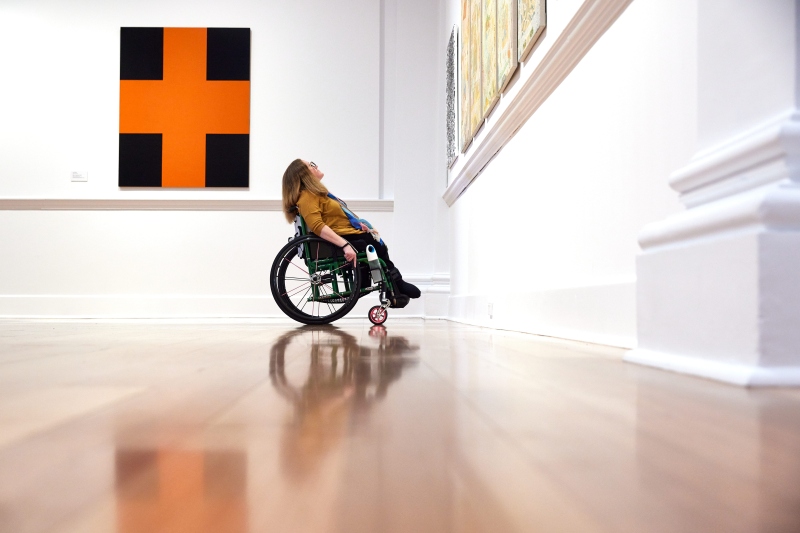 A woman in a wheelchair leans back to study a large orange and black artwork in a bright gallery at Geelong.