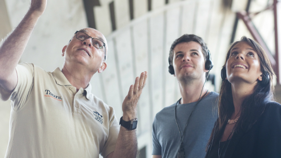 A Sydney Opera House guide gestures upward mid-explanation while two visitors wearing tour headsets look up at the building's interior shells.