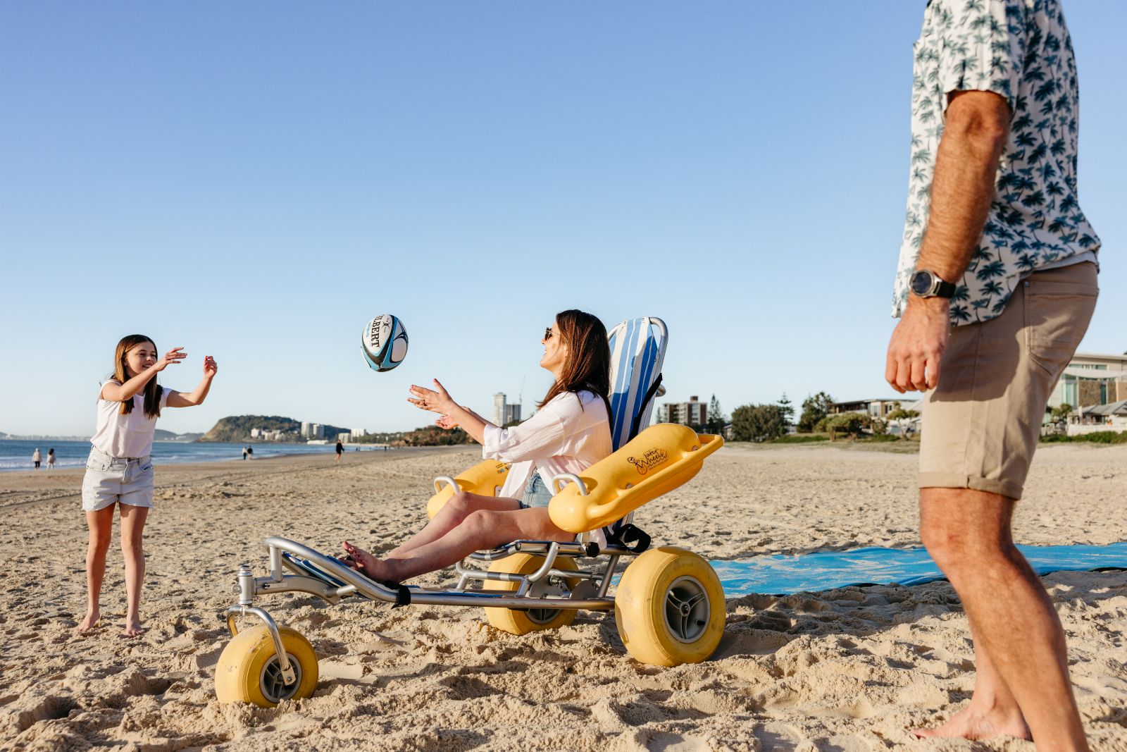 A woman seated in a yellow beach wheelchair with balloon tyres catches a football thrown by a girl on the sand, beach matting and the coastline behind them.