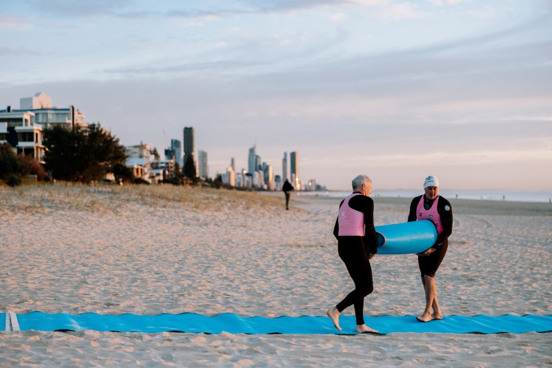 Two surfers in pink vests and wetsuits carry a blue board along beach matting at dawn, the Surfers Paradise skyline behind them.
