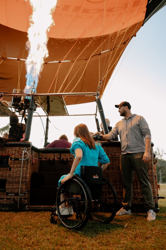 A woman in a wheelchair talks with a crew member beside a hot air balloon basket at dawn as the burner fires, inflating the orange canopy above.