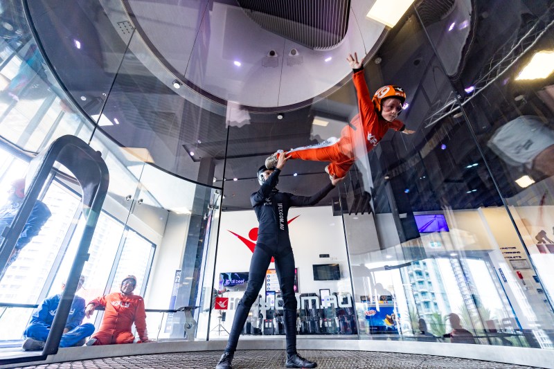 A flyer in an orange suit and helmet is supported by an instructor while floating on the column of air inside the glass wind tunnel at iFLY.