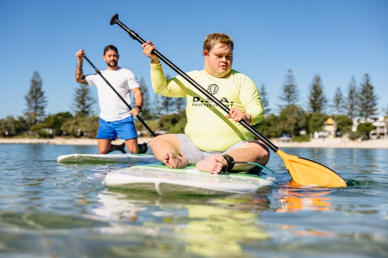 A young man with Down syndrome paddles a stand-up paddleboard from a seated position on calm water, an instructor kneeling on a board just behind him.