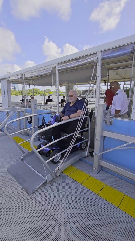 A man on a mobility scooter boards the Hopo ferry down a metal gangway with handrails, crossing yellow tactile indicators as a crew member waits by the gate.