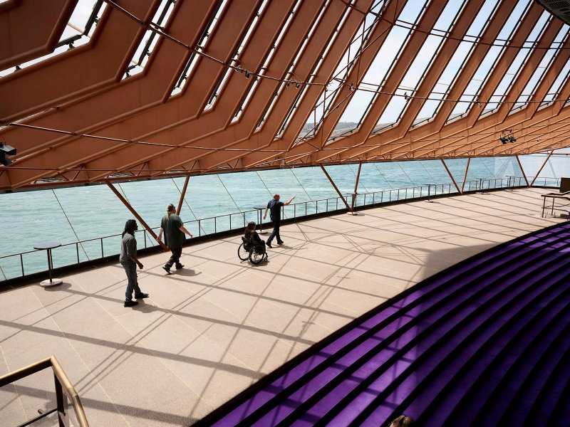 A guide gestures toward the harbour as a small tour group, including a woman using a manual wheelchair, crosses the level floor of the Sydney Opera House northern foyer beneath the building's angled glass wall.