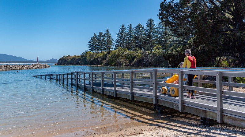 A level metal ramp with handrails runs out over a sheltered tidal inlet at Bermagui, with a yellow beach wheelchair parked at the top and a swimmer in the calm water beyond.