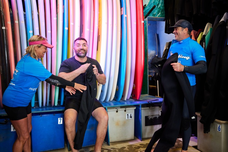 A man laughs with two Let's Go Surfing instructors as they help him into a wetsuit in the board room at Bondi, a wall of colourful surfboards behind them.