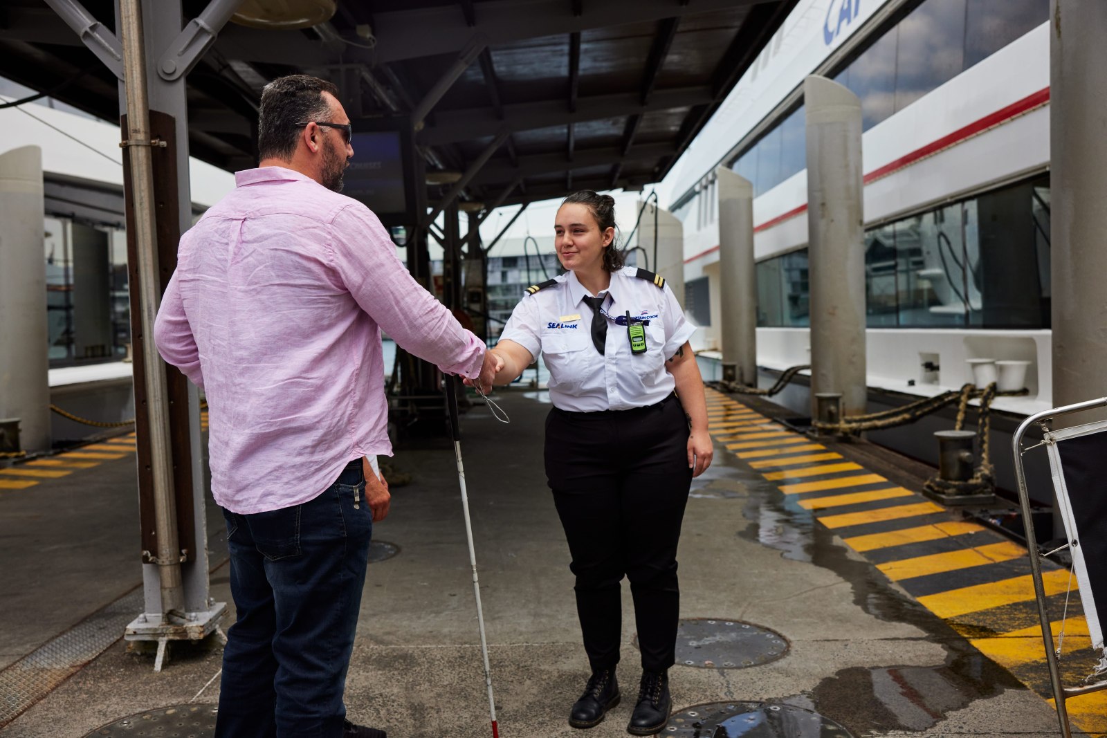 A ferry crew member greets a man with a white cane at the wharf gangway, shaking his hand and guiding him towards the vessel moored alongside.