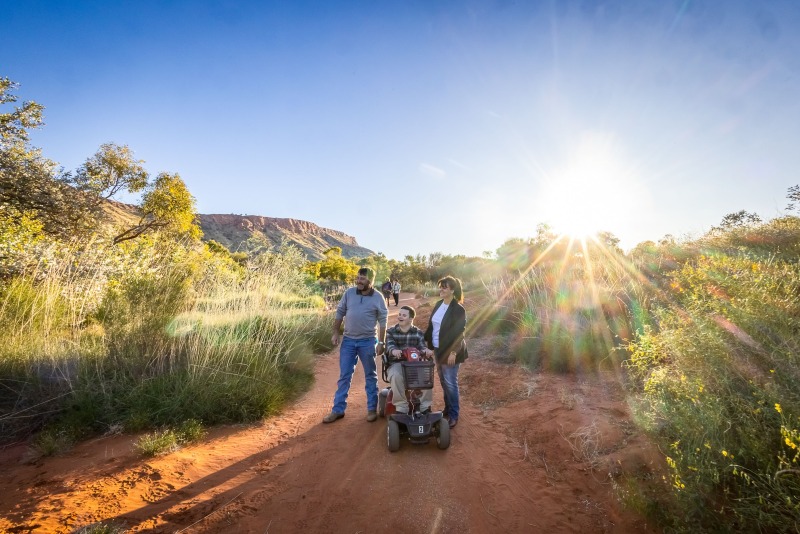 A young man on a mobility scooter travels a red dirt trail at Alice Springs Desert Park with two companions, low ranges and spinifex catching the late afternoon sun.