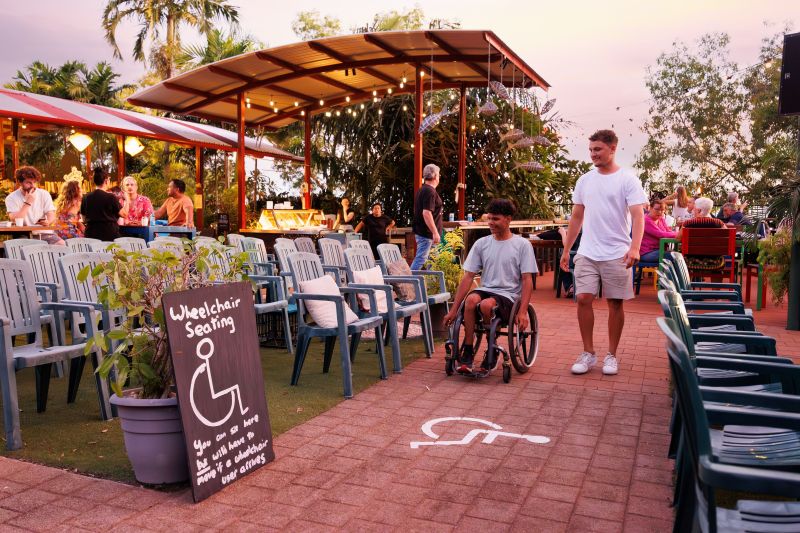 A wheelchair user arrives at the Deckchair Cinema past a chalkboard sign reading Wheelchair Seating and a wheelchair symbol painted on the level brick path, deckchairs set out under string lights at dusk.