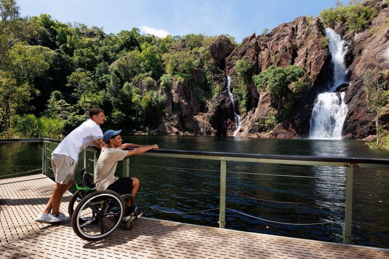 A wheelchair user and a companion look out from a level timber viewing deck across the plunge pool at Wangi Falls, twin waterfalls dropping down the escarpment opposite.