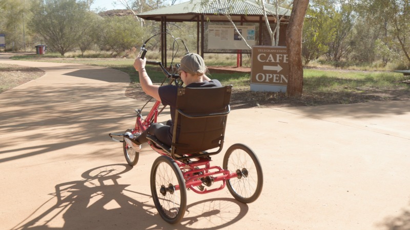 A rider on a red recumbent handcycle travels the sealed path towards the Alice Springs Telegraph Station, passing a sign for the cafe beneath river red gums.