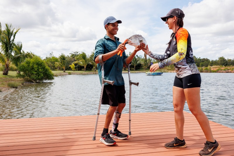 A young man using forearm crutches and a prosthetic leg grins as a guide hands him the barramundi he has just caught on the level jetty, the lake behind them.