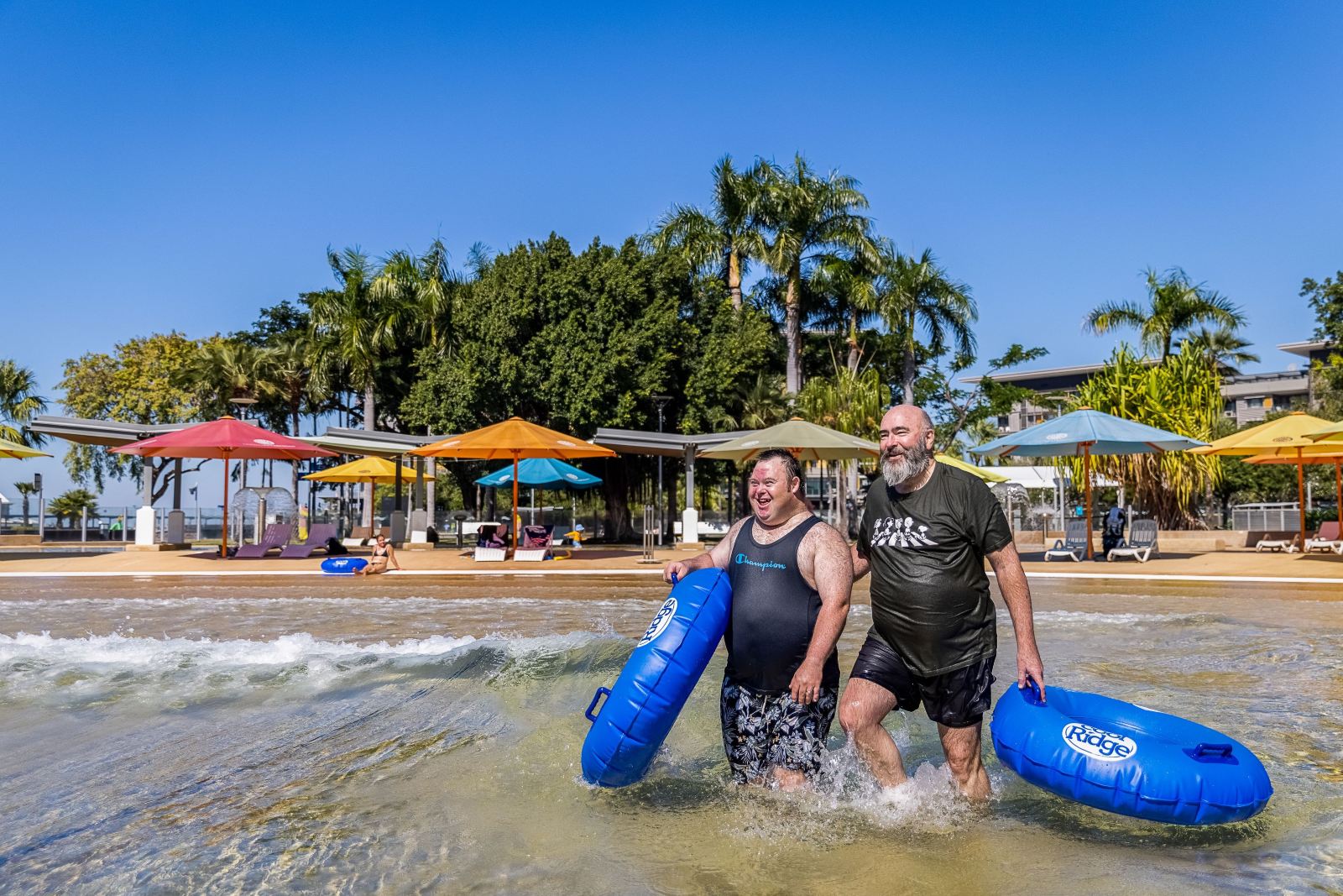Two men laugh as they wade out of the Darwin Waterfront wave lagoon carrying blue inflatable rings, palms and coloured umbrellas along the sand behind them.
