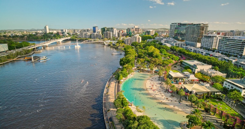 Aerial view of Streets Beach at South Bank, its turquoise lagoon and white sand ringed by palms beside the Brisbane River and city skyline.