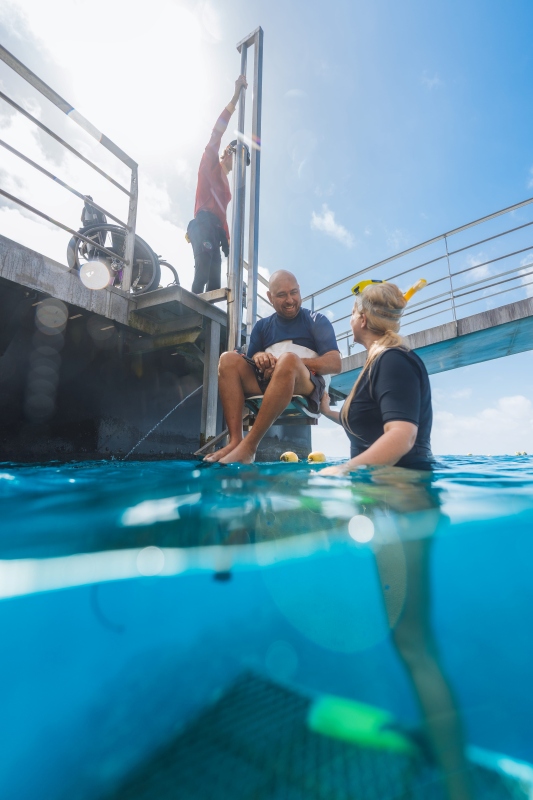 On a Great Barrier Reef pontoon, a man is lowered towards the water on an access hoist seat while a snorkeller waits beside him in the turquoise sea, his wheelchair parked on the deck above.