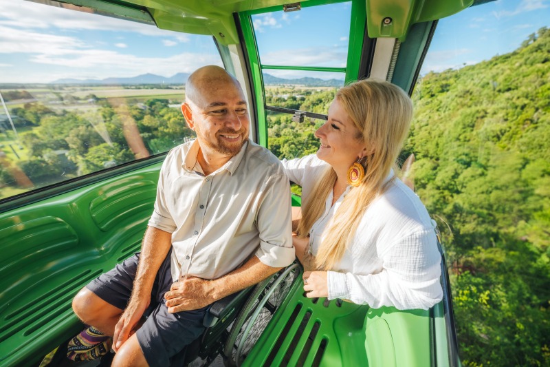 A man seated in his wheelchair and his companion smile at each other inside a green Skyrail gondola as World Heritage rainforest and the coastal plain spread out below.