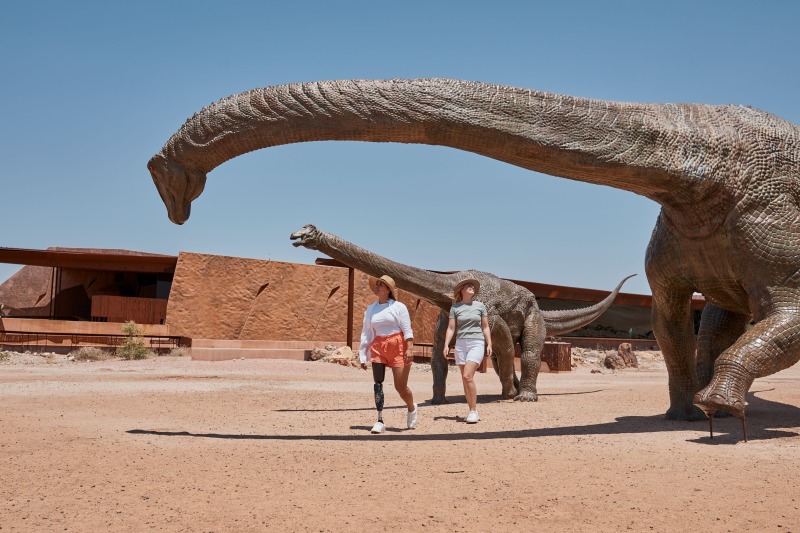Two women, one wearing a prosthetic leg, walk beneath towering life-size sauropod sculptures outside the Australian Age of Dinosaurs museum in Winton's red outback.