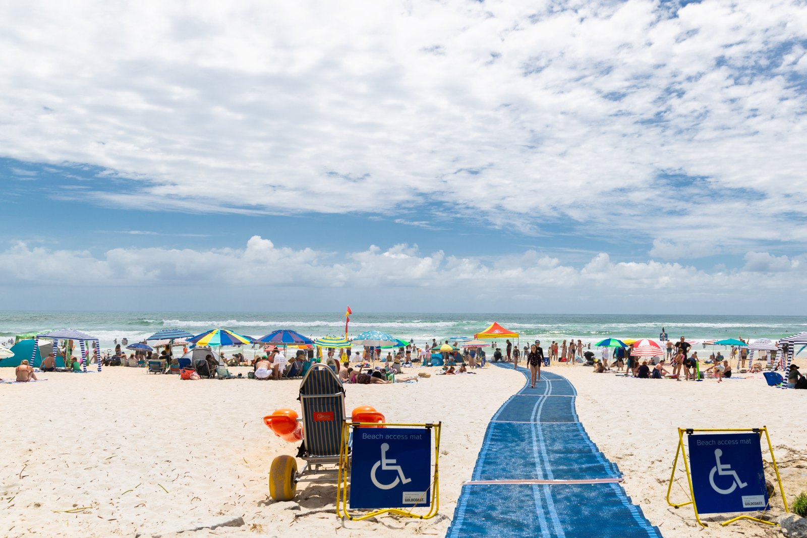 A blue beach access mat runs across golden sand towards the surf at a busy Gold Coast beach, flanked by beach access signs and a waiting beach wheelchair with balloon tyres.