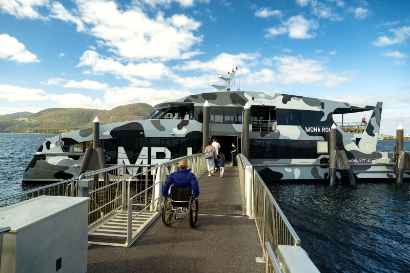 A man in a wheelchair rolls up the gangway to board the camouflage-painted MONA ROMA ferry at Berriedale, other passengers stepping aboard ahead of him.