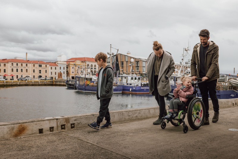 A young girl in a wheelchair laughs as her family walks with her along the Hobart waterfront, moored fishing boats and the historic H. Jones and Co building behind.