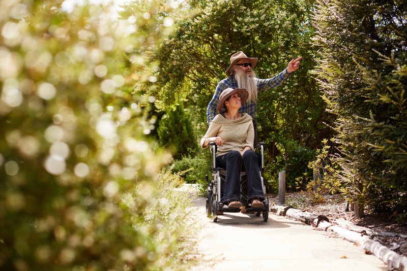 A man points out something in the trees to a woman seated in a wheelchair as they follow a sunlit sealed path through dense green garden on Bruny Island.