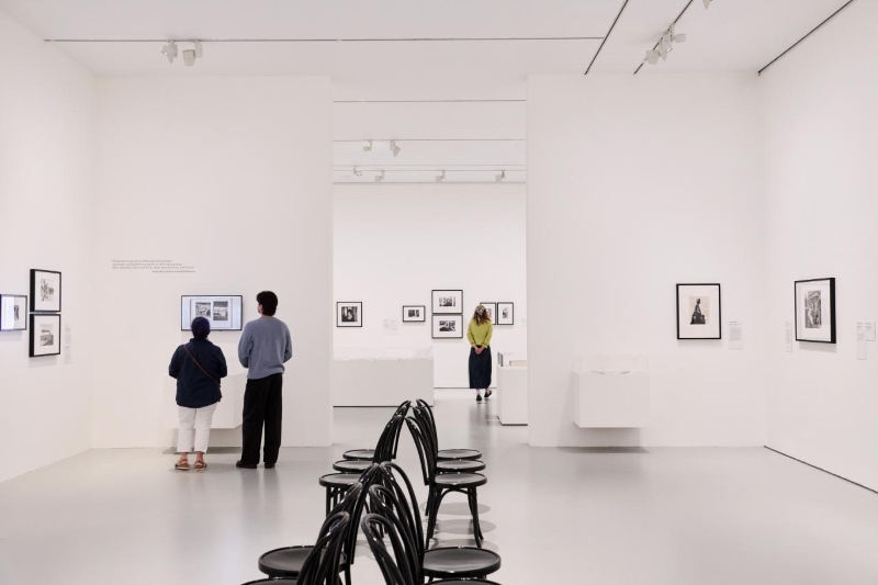 Two visitors stand quietly before framed photographs in a bright, near-empty white gallery at the NGV, with curved bentwood stools available for resting.