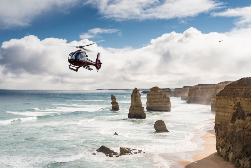 A red helicopter flies past the Twelve Apostles limestone stacks rising from turquoise surf beside the Great Ocean Road cliffs.