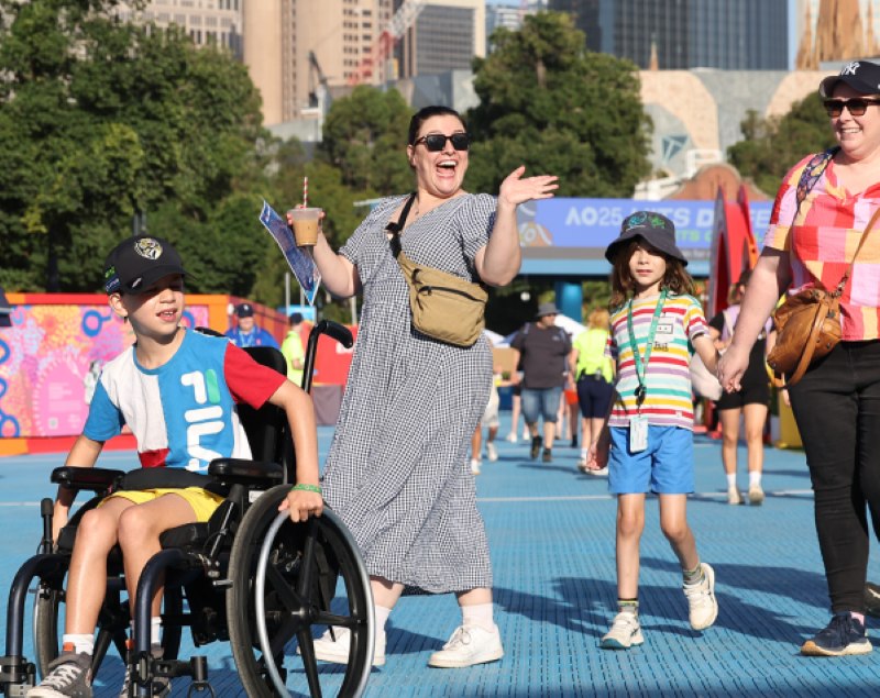 A boy using a manual wheelchair rolls along the blue level pathway at Melbourne Park with his family beside him, the city skyline behind.