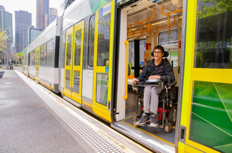 A young man in a powered wheelchair smiles as he boards a Melbourne tram through the wide accessible entry doorway, level with the raised platform stop.